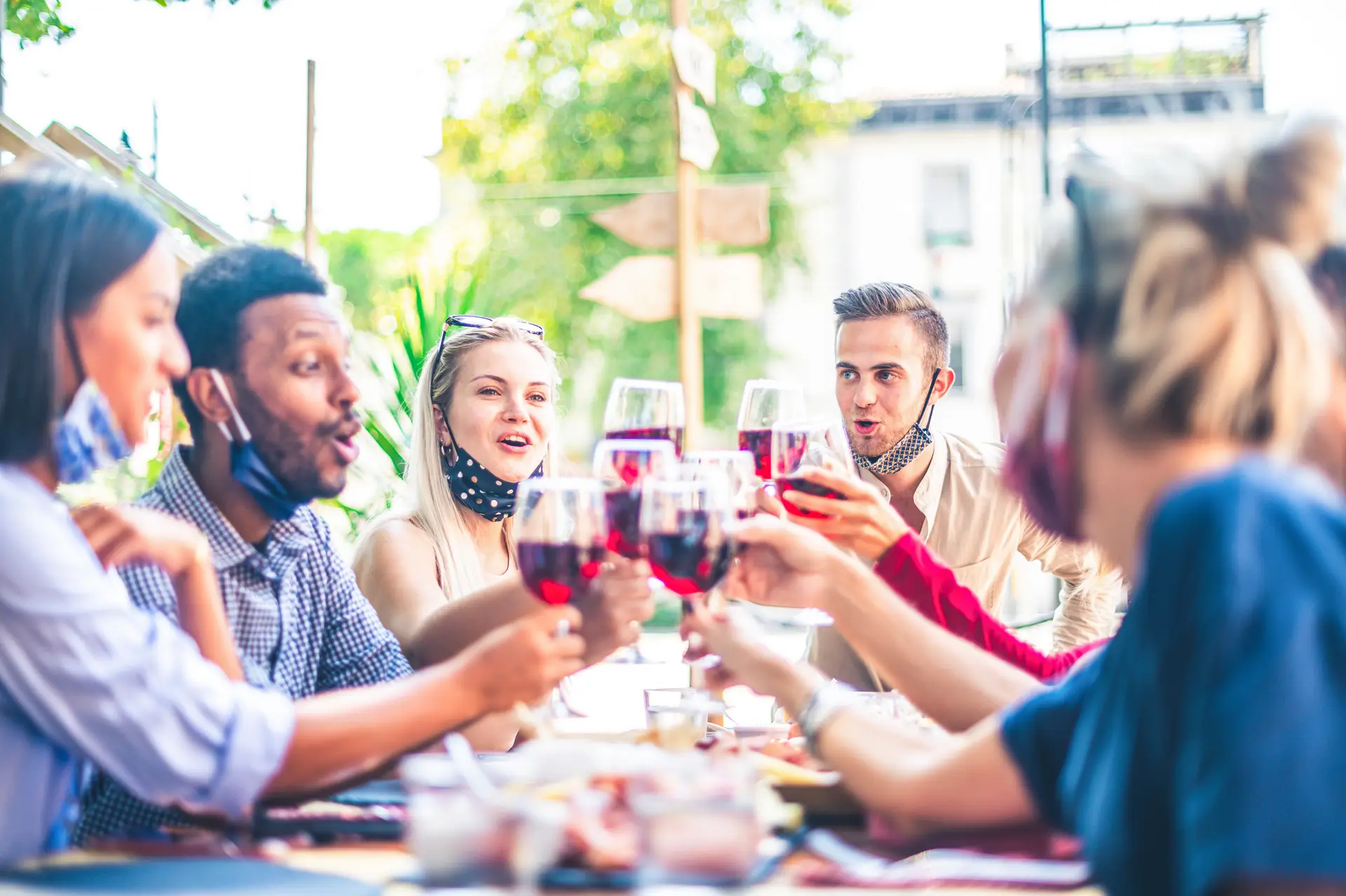 Friends toasting red wine at outdoor restaurant bar with face mask down - New normal lifestyle concept with happy people having fun together - Focus on guys behind